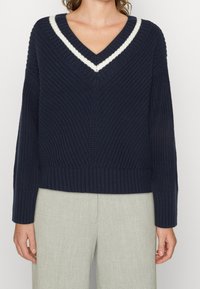 Granatowy sweter typu v-neck z teksturowanym wzorem, z białymi paskami na kołnierzu oraz ściągaczami przy mankietach i dolnej krawędzi.
