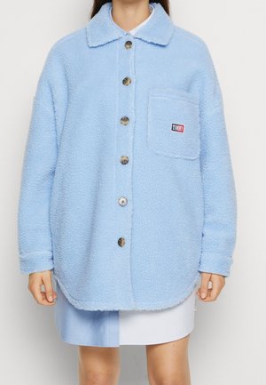 Person iført en lyseblå, blød knappet jakke med en brystlomme, der viser en lille "Tommy" logopatch.