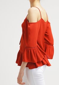 Haut rouge à épaules dénudées avec des manches évasées et un tissu texturé. Présente un ourlet évasé et des bretelles fines, associé à un pantalon blanc.