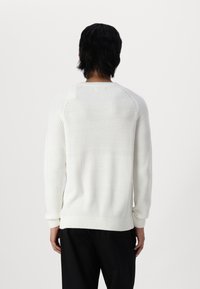 Maglione a coste color crema, maniche lunghe, scollatura tonda, texture morbida, dettagli delle cuciture visibili, abbinato a pantaloni scuri, visto da dietro.