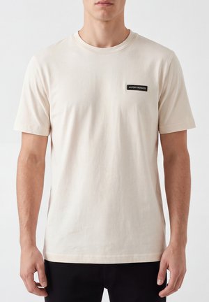 Uomo che indossa una t-shirt beige chiaro semplice, con scollo rotondo, maniche corte e una piccola toppa rettangolare nera con logo sul petto sinistro.