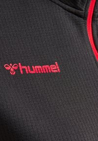 Tejido negro texturizado con rayas verticales, que presenta una cremallera roja y el logo rojo de "hummel". Primer plano del detalle y la costura de la prenda.