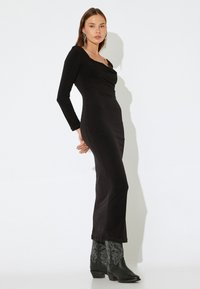 Schwarzes Kleid mit langen Ärmeln, drapiertem Ausschnitt und taillierter Silhouette. Getragen mit schwarzen, verzierten Cowboy-Stiefeln. Einfacher, strukturierter Stoff.