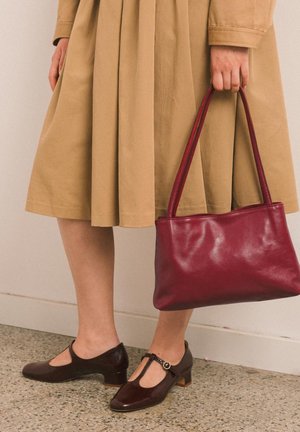 Personne portant des chaussures à brides marron et une jupe beige, tenant un sac à main en cuir rouge foncé par ses poignées.