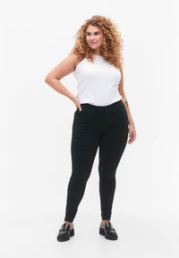 Černé skinny džíny z elastického materiálu, s vysokým pasem a předními kapsami, v kombinaci s bílým topem bez rukávů a černými mokasíny.