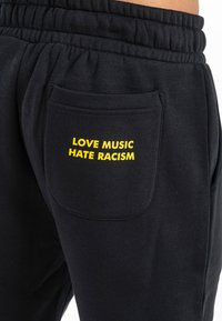 Pantalones de chándal negros con cintura elástica y bolsillos laterales. Con texto amarillo: "AMOR A LA MÚSICA ODIO AL RACISMO" en el bolsillo trasero. Tela suave.