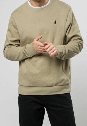 Mężczyzna noszący beżową bluzę z długim rękawem z małym czarnym logo na klatce piersiowej, poprawiający rękawy, w zestawieniu z czarnymi spodniami na jednolitym tle.