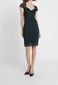 Tmavě zelenomodré krajkové šaty se čtvercovým výstřihem a krátkými krajkovými rukávy. Vlnitý lem a přiléhavý střih, doplněné černými špičatými podpatky.