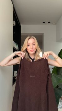 Donna bionda che tiene in mano un maglione di lana marrone scuro all'interno, in piedi davanti a uno scaffale nero e una pianta verde.