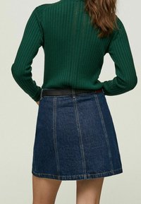 Femme portant un pull côtelé vert rentré dans une mini-jupe en denim bleu foncé avec des coutures verticales et une ceinture noire, se tenant les mains sur les hanches.