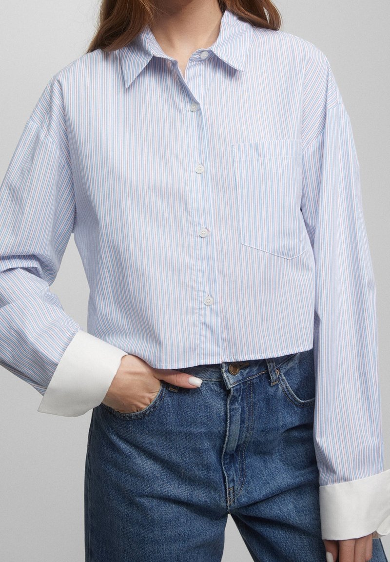 Donna che indossa una camicia cropped a righe azzurro chiaro con polsini bianchi e jeans blu a vita alta, con una mano in tasca.