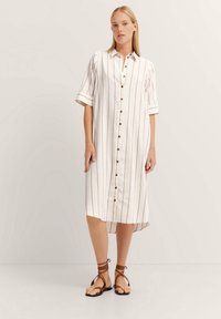 Weißes Leinenhemdkleid mit vertikalen braunen Streifen, kurzen aufgerollten Ärmeln und Knopfleiste. Kombiniert mit braunen flachen Sandalen.