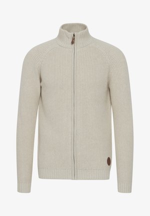 Suéter de punto beige claro con cierre, cuello alto, puños de canalé y un parche redondo marrón en la parte inferior izquierda.