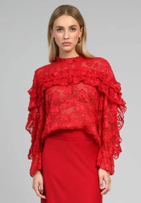 Blouse rouge transparente avec des manches longues à volants et une broderie florale. Présente un col haut et une longueur cropped, associée à une jupe rouge.