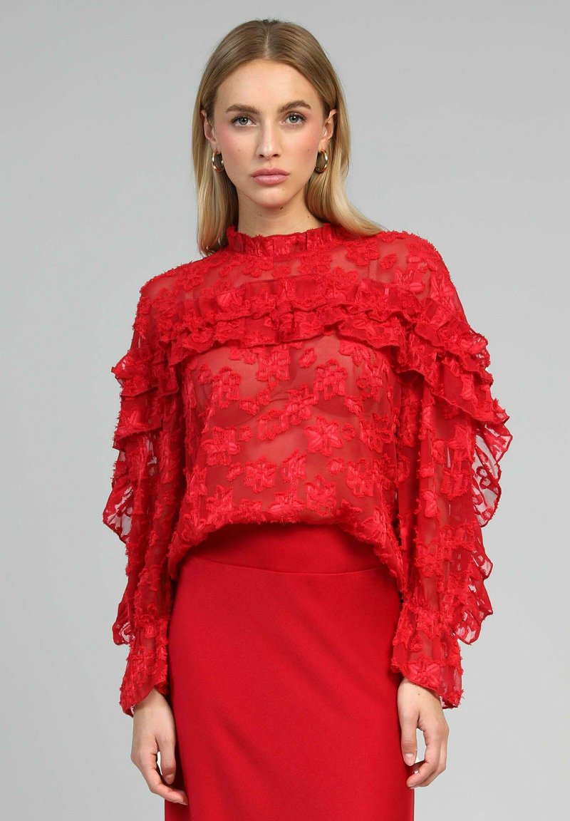 Blouse rouge transparente avec des manches longues à volants et une broderie florale. Présente un col haut et une longueur cropped, associée à une jupe rouge.