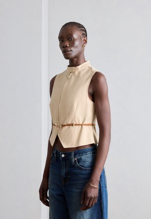 Žena s vrkočom nosí béžový bezrukávový top so zipsom a tenkým opaskom a voľné modré rifle, stojaca pred jednoduchou svetlou stenou.