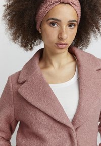 Rosa ullblandad kappa med texturerad yta och breda slag, kombinerad med ett stickat pannband. Har en vit topp under.