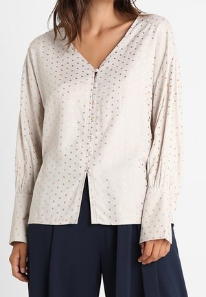 Beige Bluse mit V-Ausschnitt, langen Ärmeln und Knopfleiste. Verfügt über kleine goldene Punkte und einen Schlitz am vorderen Saum. Weiche Textur.