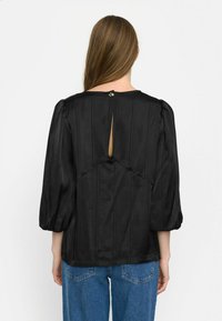Blusa nera in satin con maniche a sbuffo. Presenta righe verticali, un piccolo apertura a chiave sul retro e un bottone decorativo. Abbinata a jeans blu.