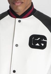 Mand iført en struktureret hvid og sort varsityjakke med maroon-stribed ribbet krave og sort patchlogo på brystet.