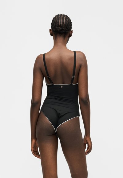 Γυναίκα με πλεξούδες στα μαλλιά, ντυμένη με μαύρο ολόσωμο μαγιό με ρυθμιζόμενους ιμάντες, στέκεται με την πλάτη προς τον θεατή σε απλό φωτεινό φόντο.