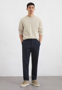 Beige Sweatshirt mit langen Ärmeln, dunkelblaue Hose und beige Sneakers. Modell steht mit Händen in den Taschen vor einem neutralen Hintergrund.