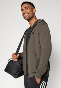 Sudadera de cremallera verde oliva con detalles negros, bolsillos delanteros y un diseño moderno, acompañada de un bolso de viaje negro.