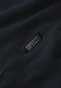 Donker marineblauwe stof met een klein zwart geweven label erop gestikt, met de merknaam "Superdry" in witte hoofdletters.
