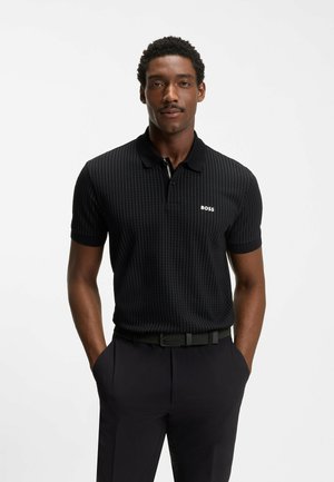 Uomo che indossa una polo nera con trama e logo "BOSS" e pantaloni scuri, con le mani in tasca, in piedi davanti a uno sfondo bianco.