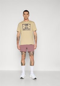 Camiseta beige con un estampado gráfico, combinada con pantalones cortos de color rosa. Zapatillas blancas y calcetines tipo barco completan el look casual.