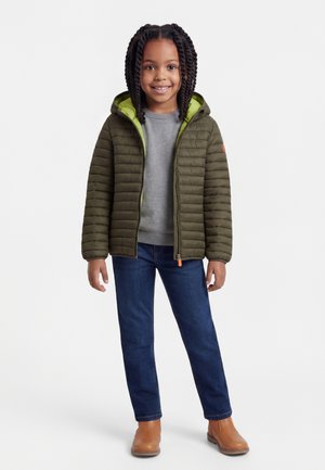 Smilende pige med flettet hår iført grøn hættejakke, grå sweater, blå jeans og beige ankelstøvler, stående på hvid baggrund.