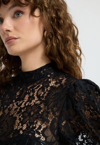 Sort blondel med høj hals, pufskuldre og indviklede blomster mønstre, med gennemsigtig tekstur og elegant design.