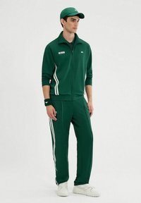 Chándal verde hecho de tela con rayas blancas a lo largo de las mangas y piernas. Complementado con una gorra y una muñequera a juego. Zapatillas blancas.