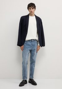 Marineblazer über einem weißen Pullover, kombiniert mit hellblauen Jeans und schwarzen Mokassins. Geschmeidige Textur, maßgeschneiderte Passform und lässiger Stil.