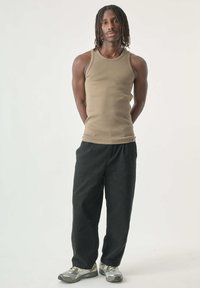 Braunes ärmelloses Tanktop, locker sitzende schwarze Hose und graue Sportschuhe mit gelben Akzenten. Das Model steht mit den Händen hinter dem Rücken vor einem schlichten Hintergrund.