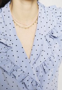 Blusa azzurro chiaro con pois neri, caratterizzata da un bordo arricciato lungo il collo. Due collane sovrapposte: una in filo rosa e una in catena dorata con ciondoli.