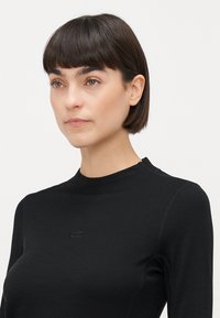 Svart langermet topp med høy hals, glatt tekstur, subtil logo på brystet, og flate sømmer for en tettsittende design.