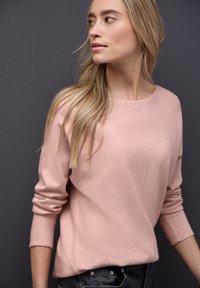 Suéter rosa claro de manga larga, con cuello redondo y acabado texturizado, combinado con jeans oscuros; detalles de costura sutil a lo largo del frente.