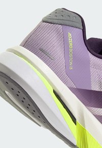 Sapatilha de corrida lilás clara com parte superior em malha, detalhes cinzentos e sola amarelo vivo. Apresenta padrão texturizado e calcanhar reforçado.