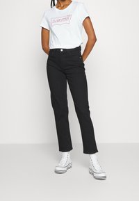 Svarta högmidjade jeans med rak passform, i kombination med en ljusblå t-shirt som har en rosa Levi's-logotyp, och vita plattformssneakers.