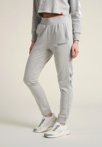 Grå sweatpants med elastisk midja, sidofickor, Hummel-logotyp och kontrasterande mörkgrå chevronmönster på sidorna.
