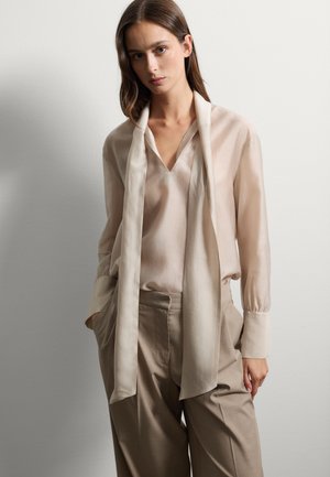 Lichtbeige blouse met een V-hals en een lange, draperende sjaal. Gecombineerd met lichtbruine, wijde broekspijpen. Glad stof en zachte textuur.