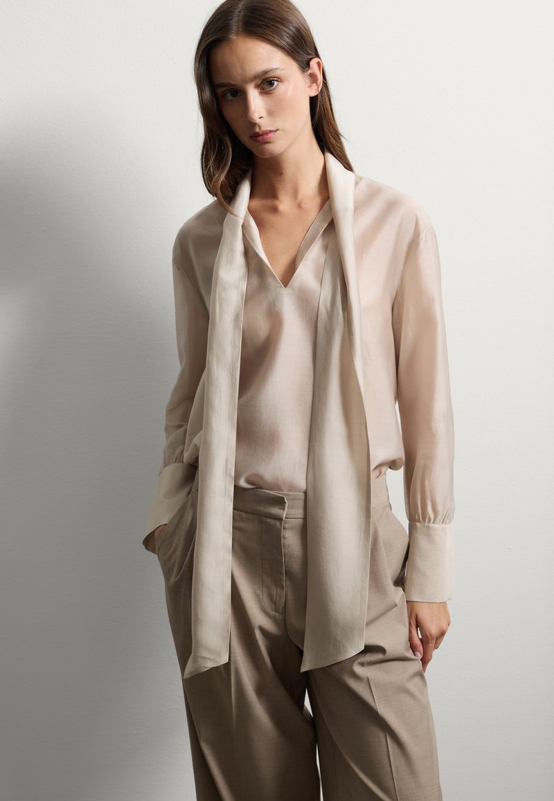 Blouse beige clair avec un décolleté en V et une longue écharpe drapée. Assortie avec un pantalon large marron clair. Tissu lisse et texture douce.