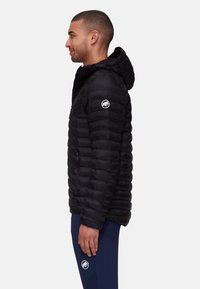 Black puffer jacket with a hood, featuring quilted sections and a logo patch on the sleeve, worn over blue pants. Smooth texture and lightweight design.