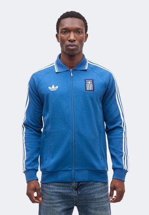 Mann trägt eine blaue Adidas Zip-Up-Trainingsjacke mit weißen Streifen und einem griechischen Emblem, kombiniert mit blauen Jeans, steht vor einem weißen Hintergrund.
