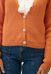 Orange strikjakke med V-hals, der har fire dekorative knapper, ribbede kanter og ærmer, parret med mørkeblå tætsiddende jeans.
