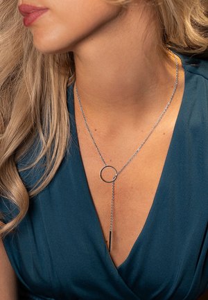 Gros plan sur une femme portant un collier lasso argenté avec un pendentif circulaire, vêtue d'un haut col en V vert sarcelle foncé.