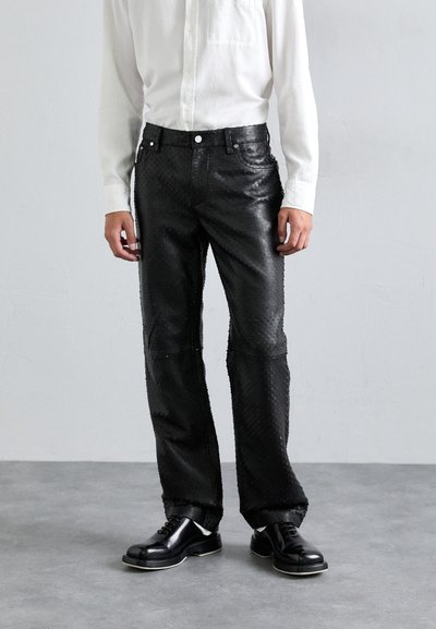 Pantaloni in pelle nera con un motivo testurizzato, design a gamba dritta e cuciture a contrasto, abbinati a una camicia bianca con bottoni e scarpe nere.