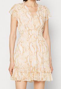 Femme portant une robe courte en imprimé serpent beige et tan avec un décolleté en V, des manches courtes, une taille élastique et un ourlet froncé sur fond uni.