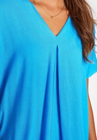 Top blu, a maniche corte in tessuto leggero, con scollo a V e dettaglio plissettato davanti, texture liscia e vestibilità rilassata.